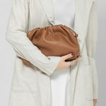 Een vrouw in een lichtbeige blazer en witte jurk houdt de Serena Zachte Leren Pouch Tas van Liora The Label - een minimalistische, moderne, soepele tas - voor haar bovenlichaam. Haar donkere schouderlange haar en zilveren ring maken haar look compleet.