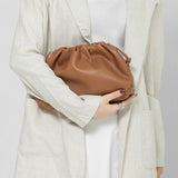 Een vrouw in een lichtbeige blazer en witte jurk houdt de Serena Zachte Leren Pouch Tas van Liora The Label - een minimalistische, moderne, soepele tas - voor haar bovenlichaam. Haar donkere schouderlange haar en zilveren ring maken haar look compleet.