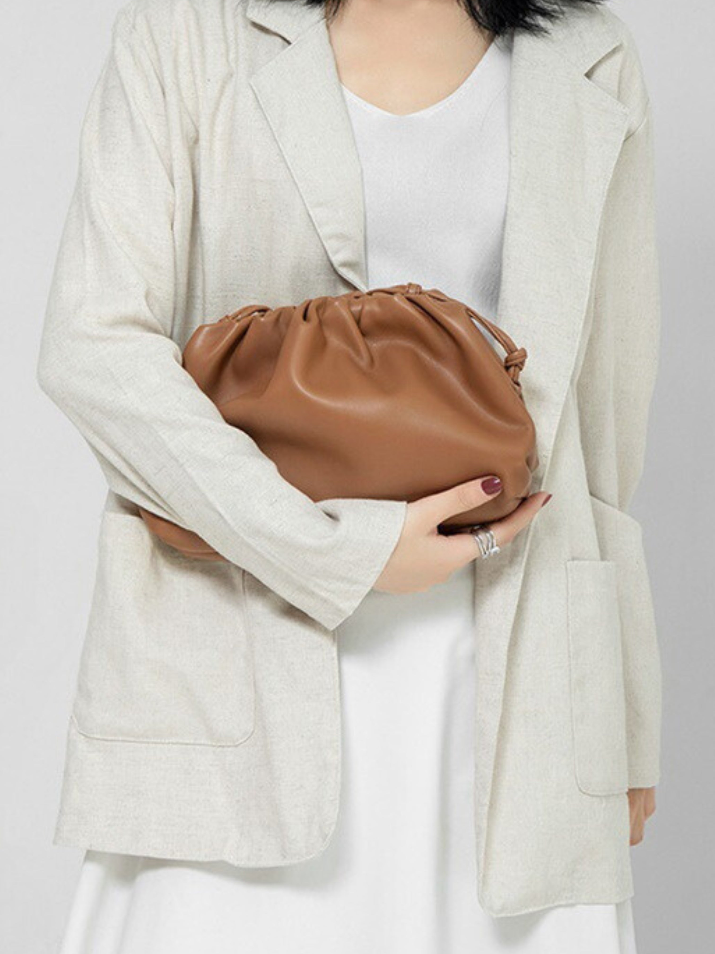Een vrouw in een lichtbeige blazer en witte jurk houdt de Serena Zachte Leren Pouch Tas van Liora The Label - een minimalistische, moderne, soepele tas - voor haar bovenlichaam. Haar donkere schouderlange haar en zilveren ring maken haar look compleet.