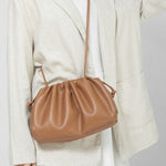 Een persoon in een lichtbeige blazer en witte jurk draagt de Serena Zachte Leren Pouch Tas van Liora The Label, met een minimalistisch, modern ontwerp met soepele leer en een lange riem. De nadruk ligt op de eenvoud van de outfit en het elegante ontwerp van de tas.