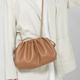 Een persoon in een lichtbeige blazer en witte jurk draagt de Serena Zachte Leren Pouch Tas van Liora The Label, met een minimalistisch, modern ontwerp met soepele leer en een lange riem. De nadruk ligt op de eenvoud van de outfit en het elegante ontwerp van de tas.