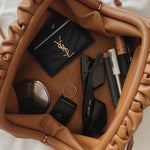 De Serena Zachte Leren Pouch Tas van Liora The Label, in bruine wollook en boterzacht leer, bevat een zwarte zonnebril, een zwarte kaarthouder met goudkleurig logo, lipproducten, een compact spiegeltje, een sleutel en een haarstrikje.