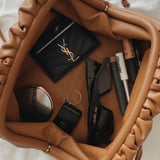 De Serena Zachte Leren Pouch Tas van Liora The Label, in bruine wollook en boterzacht leer, bevat een zwarte zonnebril, een zwarte kaarthouder met goudkleurig logo, lipproducten, een compact spiegeltje, een sleutel en een haarstrikje.