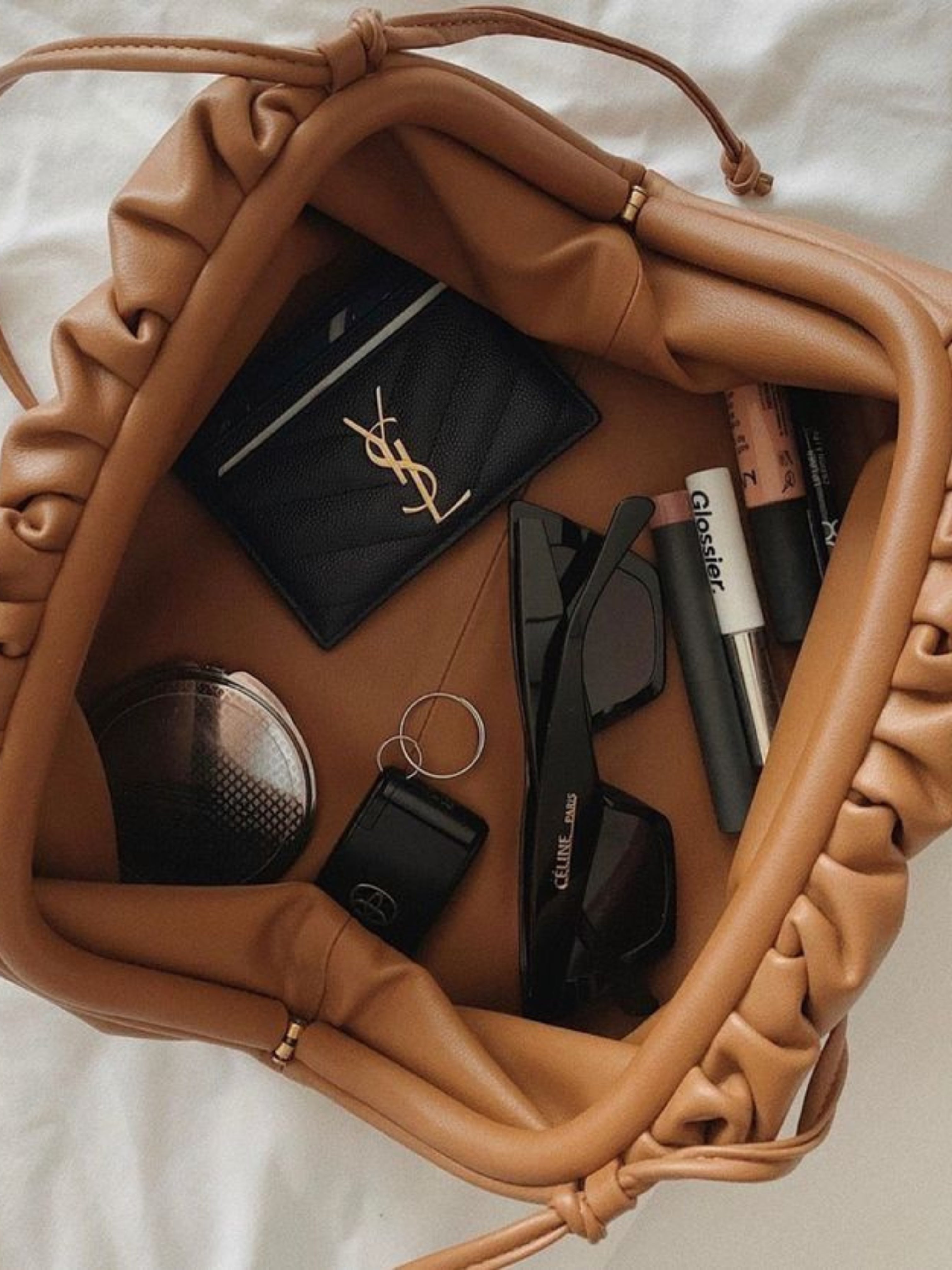 De Serena Zachte Leren Pouch Tas van Liora The Label, in bruine wollook en boterzacht leer, bevat een zwarte zonnebril, een zwarte kaarthouder met goudkleurig logo, lipproducten, een compact spiegeltje, een sleutel en een haarstrikje.