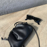 Een zwarte zonnebril en de Liora The Label Serena Zachte Leren Tas - Minimalistisch, Modern & Soepel rusten op een licht stenen oppervlak in fel zonlicht.