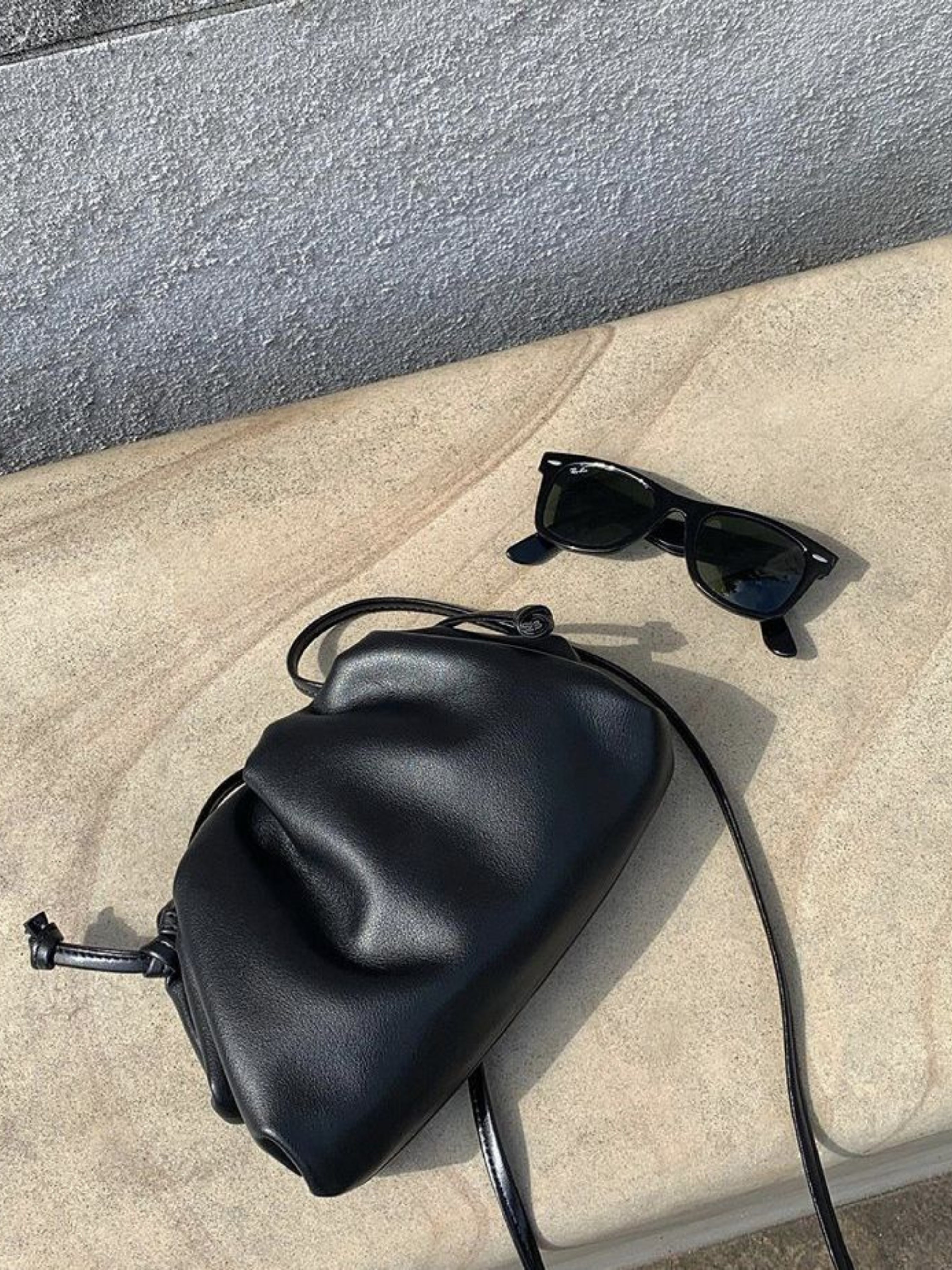 Een zwarte zonnebril en de Liora The Label Serena Zachte Leren Tas - Minimalistisch, Modern & Soepel rusten op een licht stenen oppervlak in fel zonlicht.