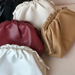 Vijf Serena Zachte Leren Pouch Tassen van Liora The Label, in zwart, wit, tan, rood en gebroken wit boterzacht leer, zijn gerangschikt op beige stof. Deze minimalistische, moderne buidels hebben een zacht ruchedesign en overlappen elkaar.