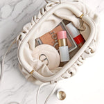 De Serena Zachte Leren Pouch Tas van Liora The Label, in crème boterzacht leer, ligt open op het marmer en onthult make-up items, een compact spiegeltje, roze lippenstift en blush. Een beige etui en een gouden munt accentueren het minimalistische moderne ontwerp.