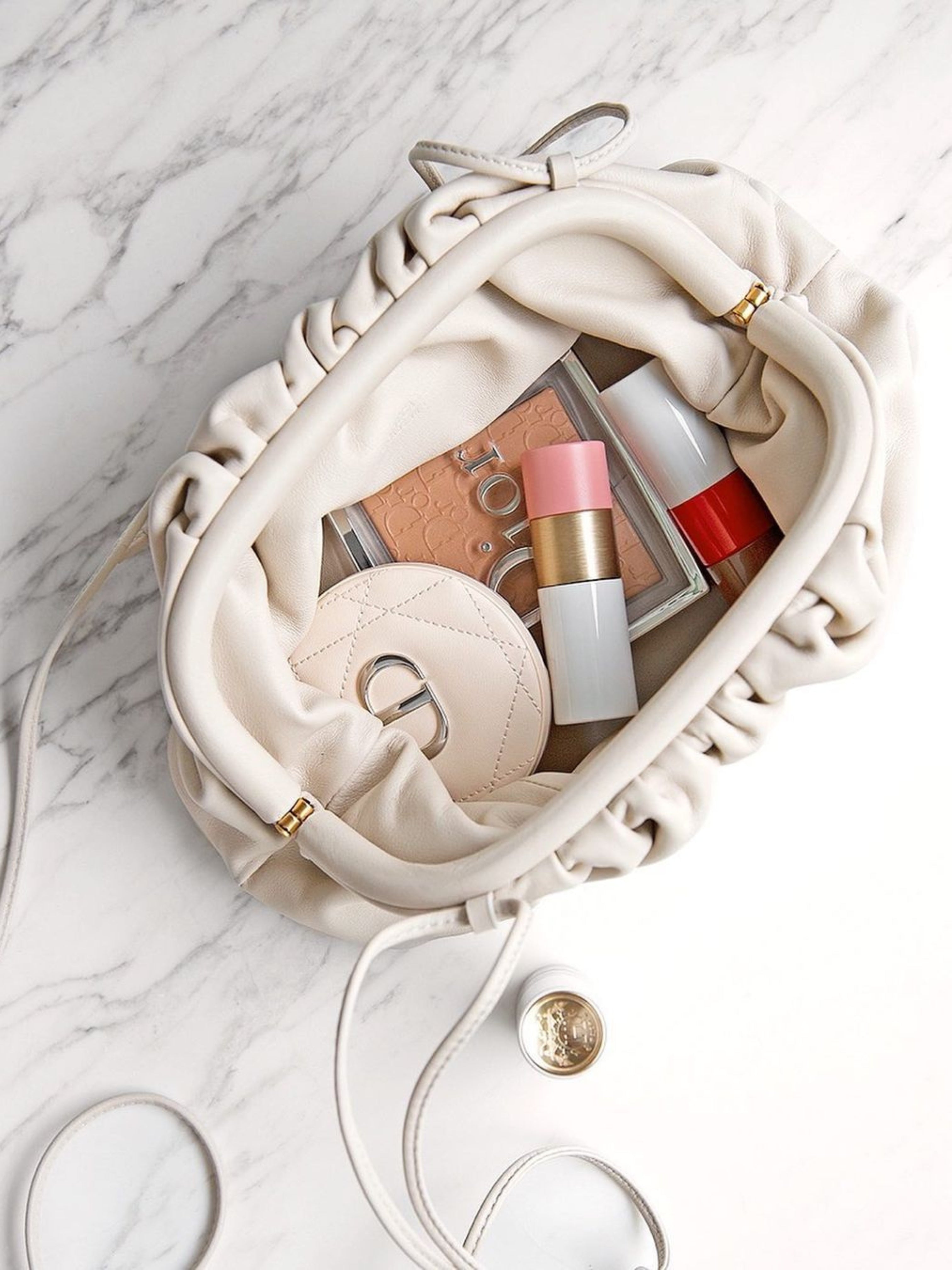 De Serena Zachte Leren Pouch Tas van Liora The Label, in crème boterzacht leer, ligt open op het marmer en onthult make-up items, een compact spiegeltje, roze lippenstift en blush. Een beige etui en een gouden munt accentueren het minimalistische moderne ontwerp.