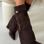 Een persoon zit in kleermakerszit en draagt Liora The Label Dames Kniehoog Laarzen met Brede Flare Bovenkant en Blokhak-chique, donkerbruine suède laarzen met spitse tenen en een metalen gesp, perfect voor een stijlvolle look.