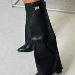 Een persoon staat op een lichte vloer en draagt Liora The Label Dames Kniehoog Laarzen met Brede Flare Bovenkant en Blokhak, met wijde, uitlopende bovenkanten die de schoenen bijna bedekken.