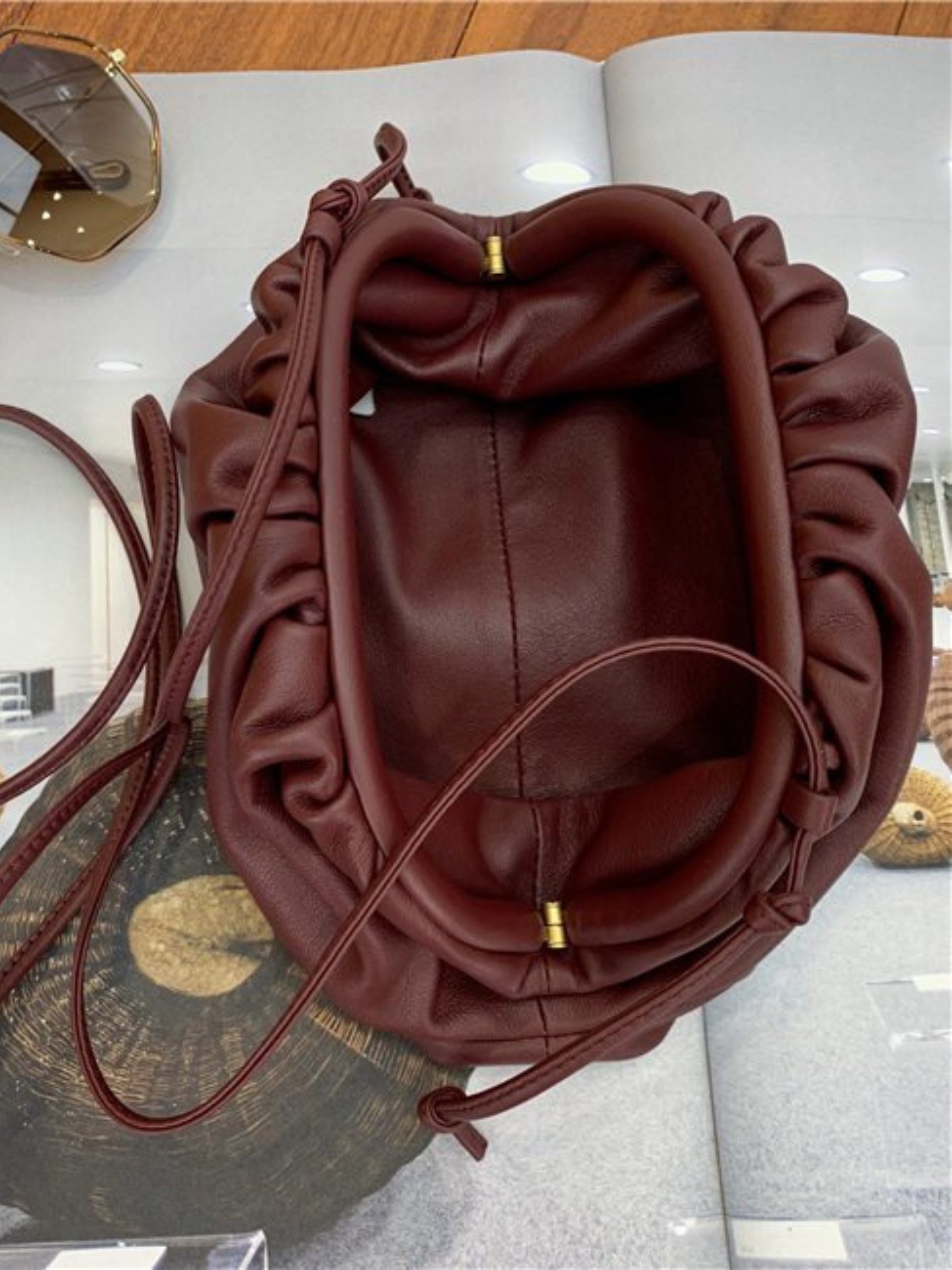 De Serena Zachte Leren Pouch Tas van Liora The Label, een kastanjebruin minimalistisch leren tasje met gouden accenten, ligt open op een tijdschrift op een houten oppervlak, met een zonnebril zichtbaar in de linkerbovenhoek.