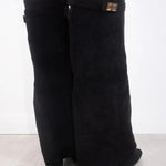 Een vrouw draagt Liora The Label Dames Kniehoog Laarzen met Brede Flare Bovenkant en Blokhak met gouden gespaccenten, staande op een licht houten vloer tegen een witte achtergrond. Deze laarzen voegen elegantie toe aan elke look.