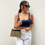 Een vrouw in een marineblauwe top, een witte broek met hoge taille en een zwarte zonnebril staat tegen een witte muur en houdt de Liora The Label Aurora Signature Mini Leren Handtas - Vrouwen Gestructureerde Top Handle Tas vast. Ze draagt gouden sieraden en heeft haar haar naar achteren getrokken.