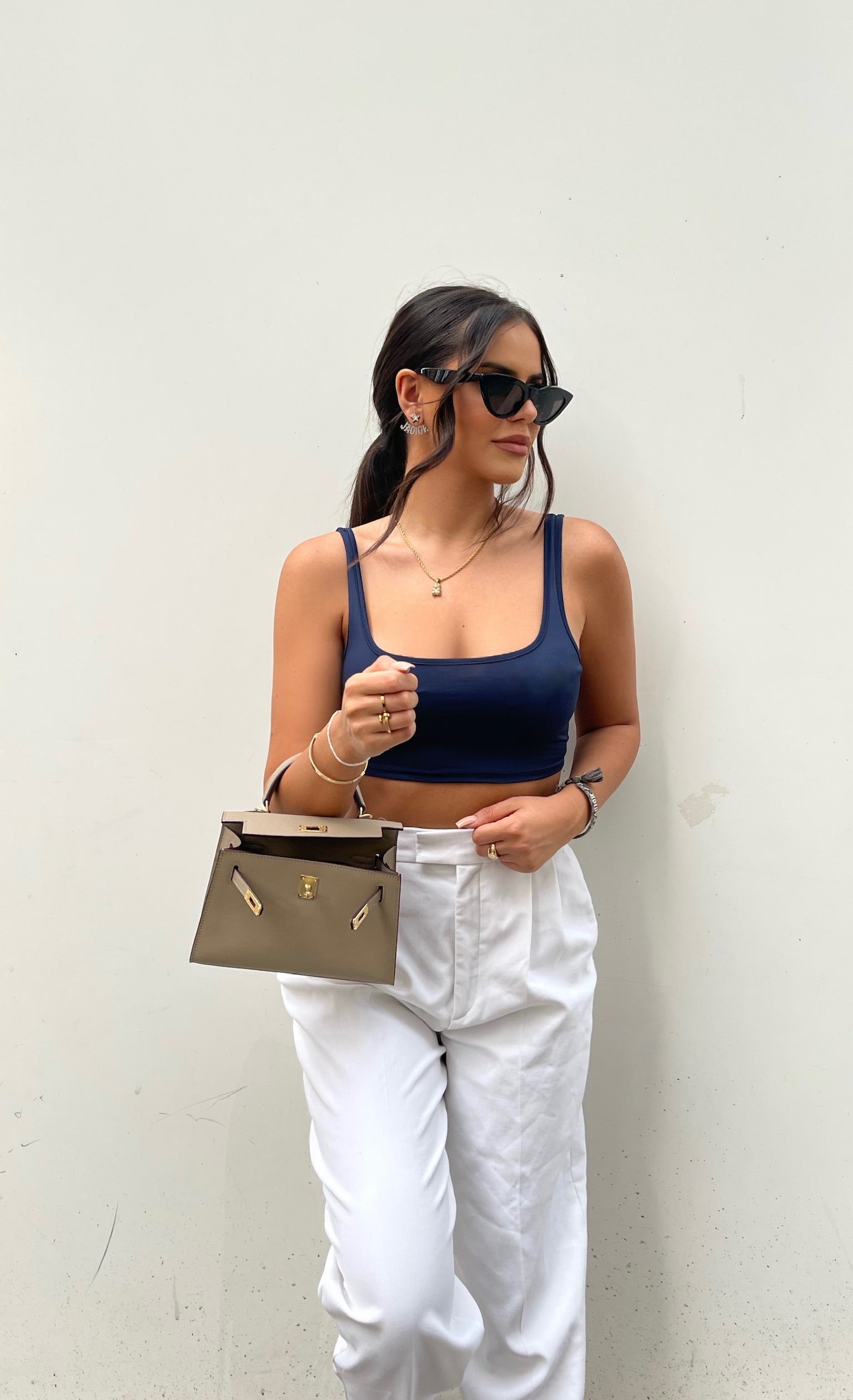 Een vrouw in een marineblauwe top, een witte broek met hoge taille en een zwarte zonnebril staat tegen een witte muur en houdt de Liora The Label Aurora Signature Mini Leren Handtas - Vrouwen Gestructureerde Top Handle Tas vast. Ze draagt gouden sieraden en heeft haar haar naar achteren getrokken.