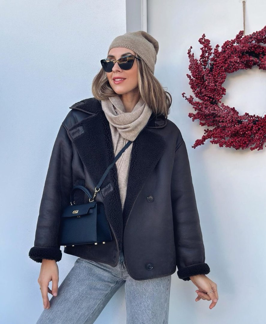 Een vrouw met zonnebril, beige muts, sjaal en zwarte omgekeerde jas staat bij een witte muur met een krans van rode bessen. Ze draagt de Liora The Label Aurora Signature Mini Leren Handtas - Vrouwen Gestructureerde Top Handle Tas.