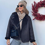 Een vrouw met zonnebril, beige muts, sjaal en zwarte omgekeerde jas staat bij een witte muur met een krans van rode bessen. Ze draagt de Liora The Label Aurora Signature Mini Leren Handtas - Vrouwen Gestructureerde Top Handle Tas.