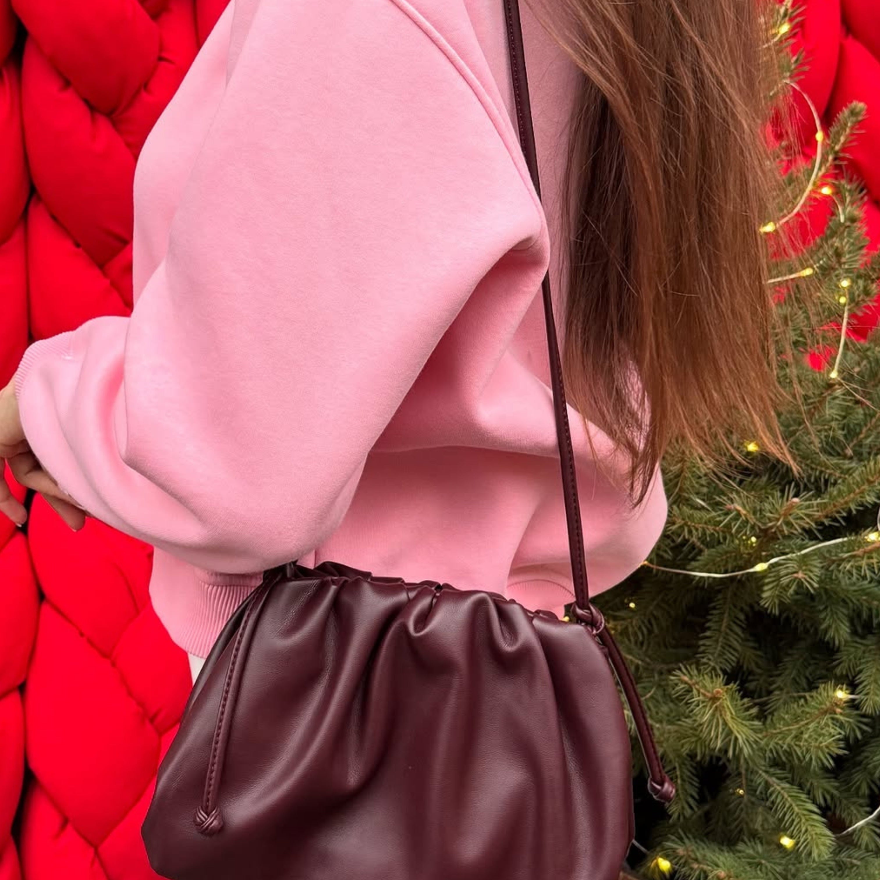 Een persoon in een roze sweater en witte broek draagt de Liora The Label Serena Zachte Leren Pouch Tas-minimalistisch, modern & soepel. De achtergrond toont een grote rode structuur en een groene dennenboom met kleine gele lichtjes.