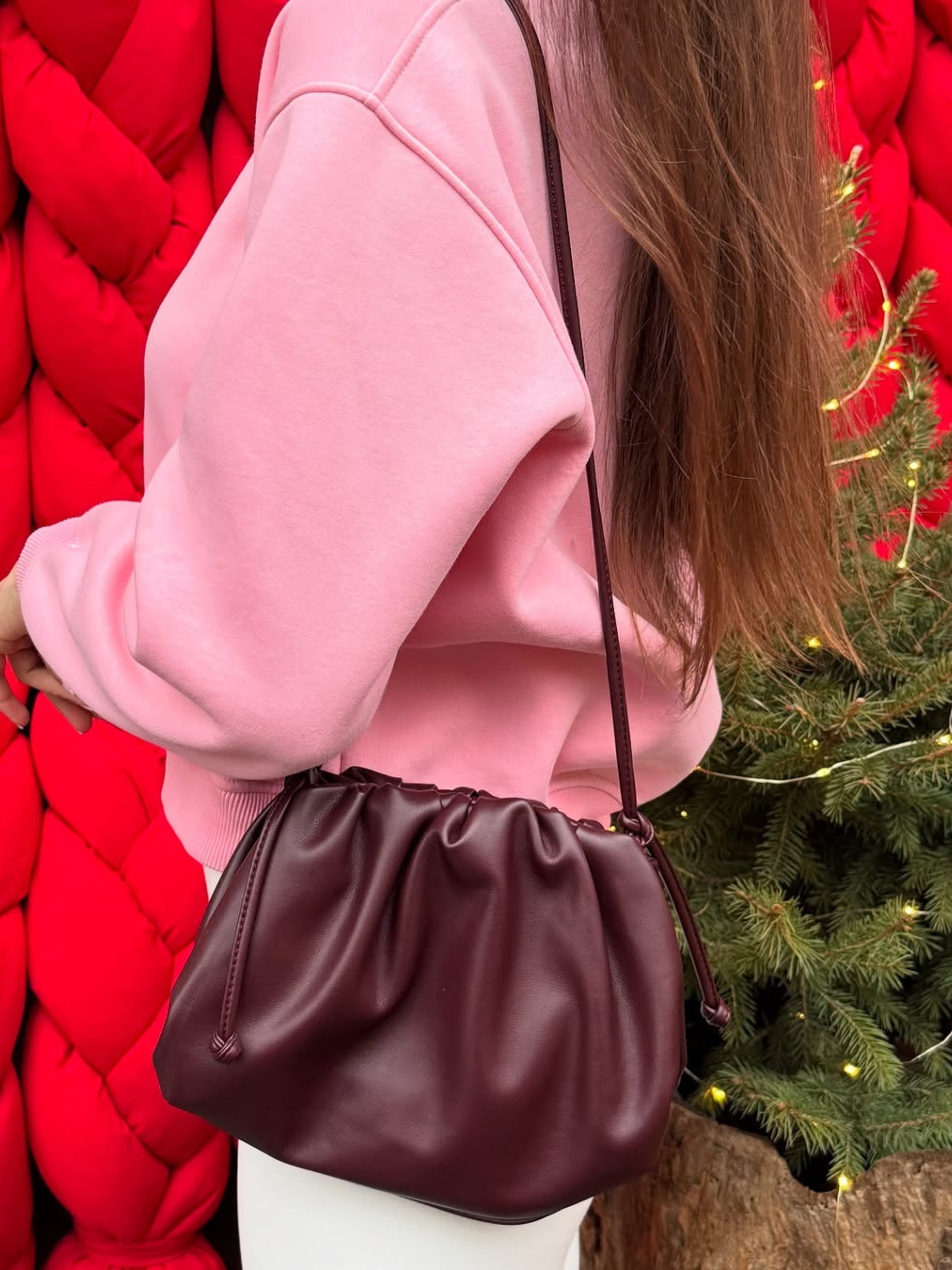 Een persoon in een roze sweater en witte broek draagt de Liora The Label Serena Zachte Leren Pouch Tas-minimalistisch, modern & soepel. De achtergrond toont een grote rode structuur en een groene dennenboom met kleine gele lichtjes.