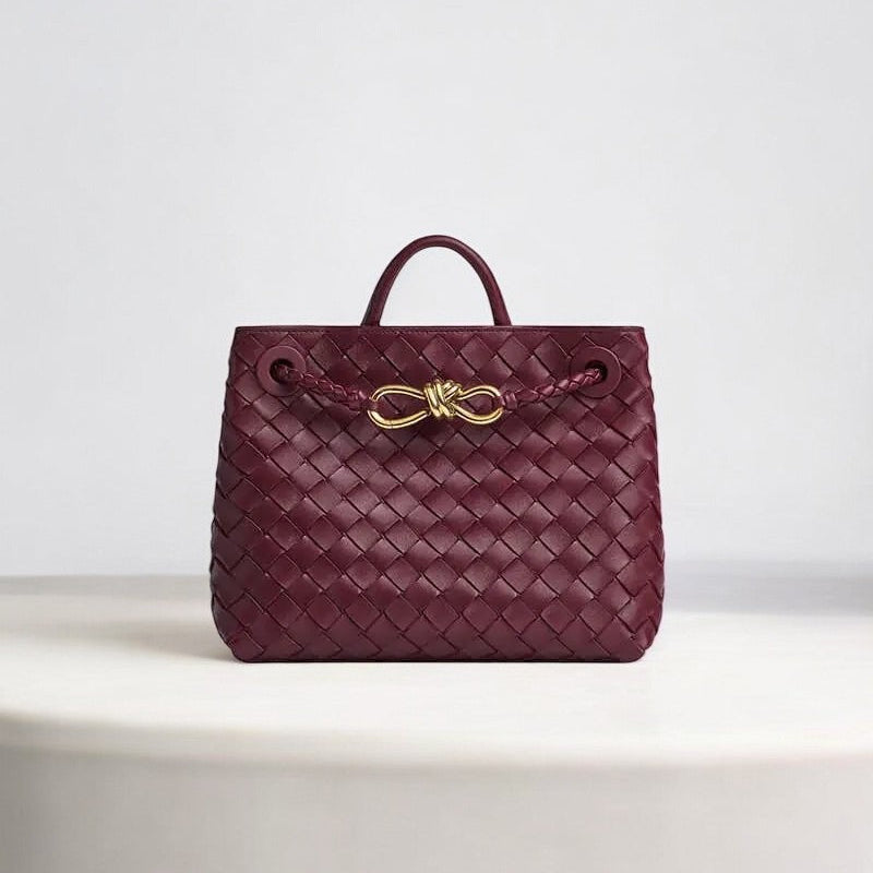 De Alyn Zwarte Geweven Midi Leren Tote Bag van Liora The Label heeft een ontwerp van geweven leer, een handvat aan de bovenkant en een gouden gesp, luxe weergegeven op een wit oppervlak met een effen lichte achtergrond.