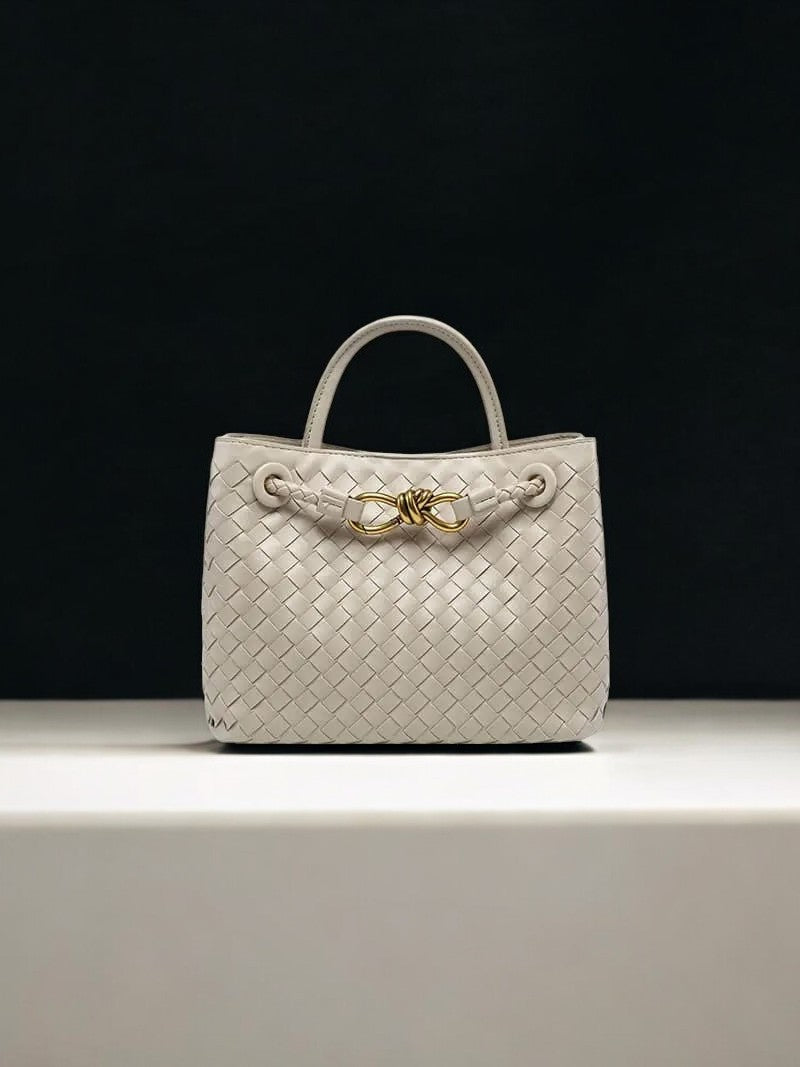 De Alyn Zwarte Geweven Midi Leren Tote Bag van Liora The Label is een moeiteloos elegante zwarte leren tas met korte hengsels en gouden knoopdetail, afgebeeld op een wit vlak tegen een zwarte achtergrond.