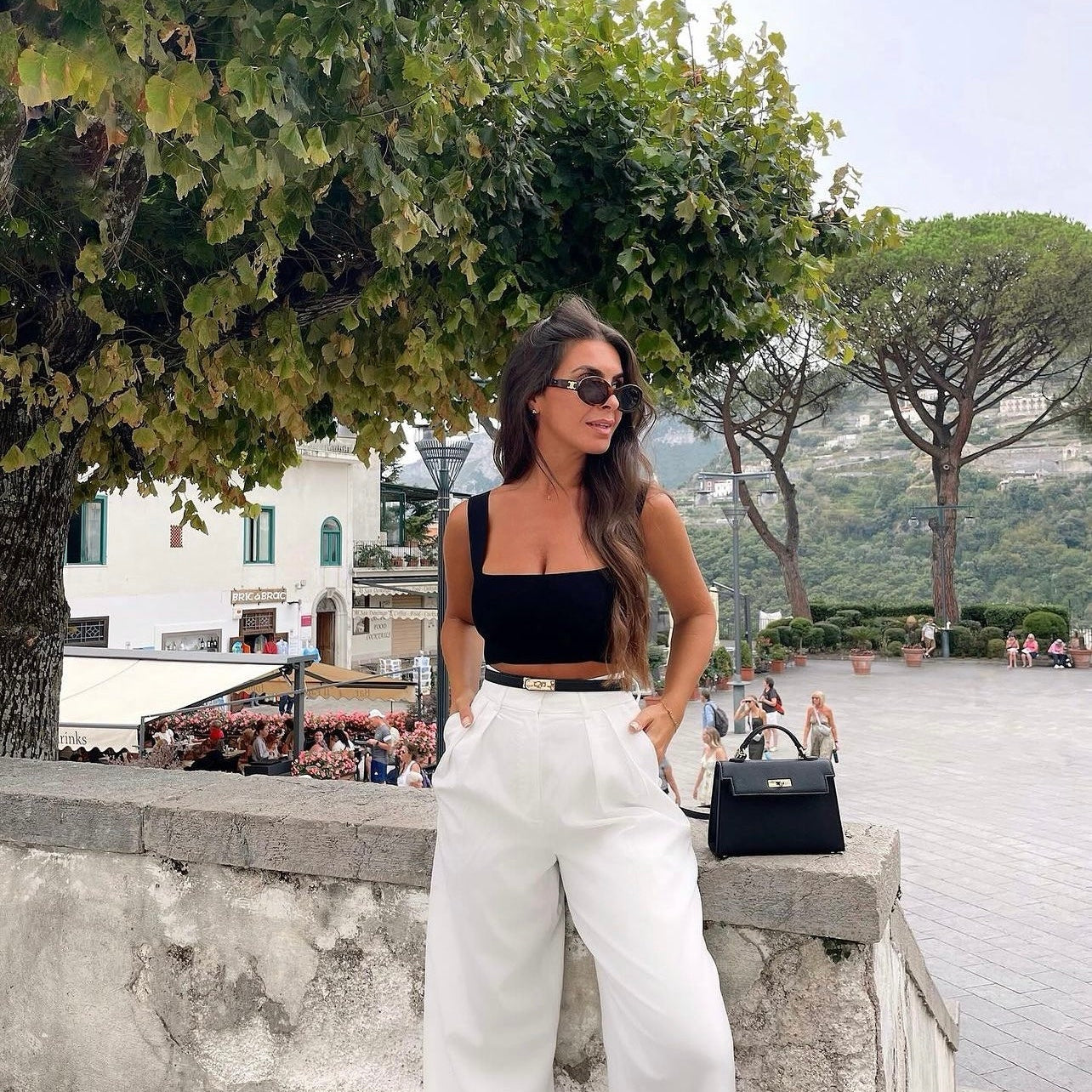 Een vrouw met lang bruin haar, een zonnebril, een zwarte top en een witte broek met hoge taille staat buiten bij een stenen muur en houdt de Liora The Label Aurora Signature Mini Leren Handtas vast. Op de achtergrond staan lommerrijke bomen en een druk plein.