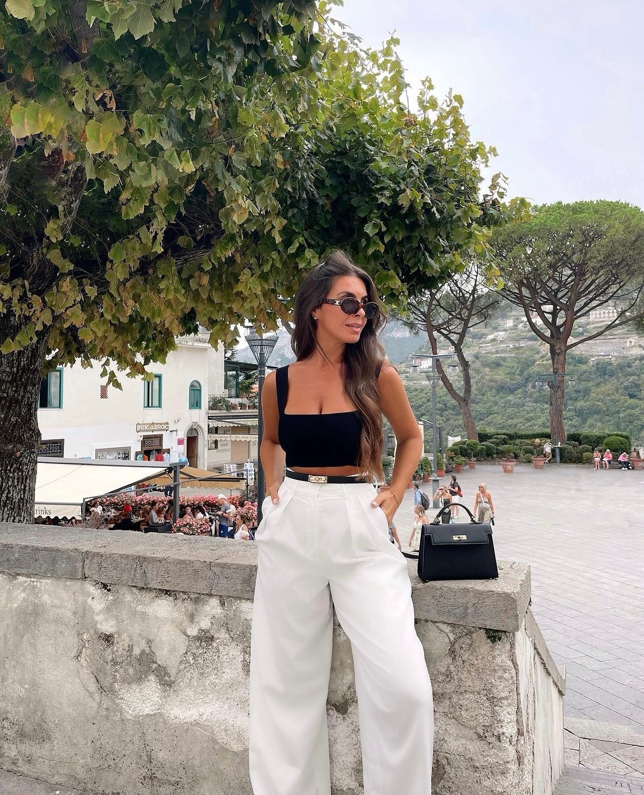 Een vrouw met lang bruin haar, een zonnebril, een zwarte top en een witte broek met hoge taille staat buiten bij een stenen muur en houdt de Liora The Label Aurora Signature Mini Leren Handtas vast. Op de achtergrond staan lommerrijke bomen en een druk plein.