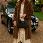Een vrouw in een lange bruine bontjas, crèmekleurige jurk en zwarte laarzen staat op een oprit met grind en houdt de Galatea Elegante Zwarte Leren Handtas van Liora The Label vast, met een klassieke zwarte auto en bakstenen huis op de achtergrond.