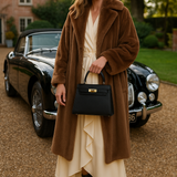 Een vrouw in een lange bruine bontjas, crèmekleurige jurk en zwarte laarzen staat op een oprit met grind en houdt de Galatea Elegante Zwarte Leren Handtas van Liora The Label vast, met een klassieke zwarte auto en bakstenen huis op de achtergrond.