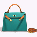 De Jovienne Luxe Teal & Tan Leren Handtas van Liora The Label heeft een handvat aan de bovenkant, contrasterende banden van bruin leer, goudkleurige hardware en een bijpassende afneembare schouderriem, afgebeeld op een lichte achtergrond.