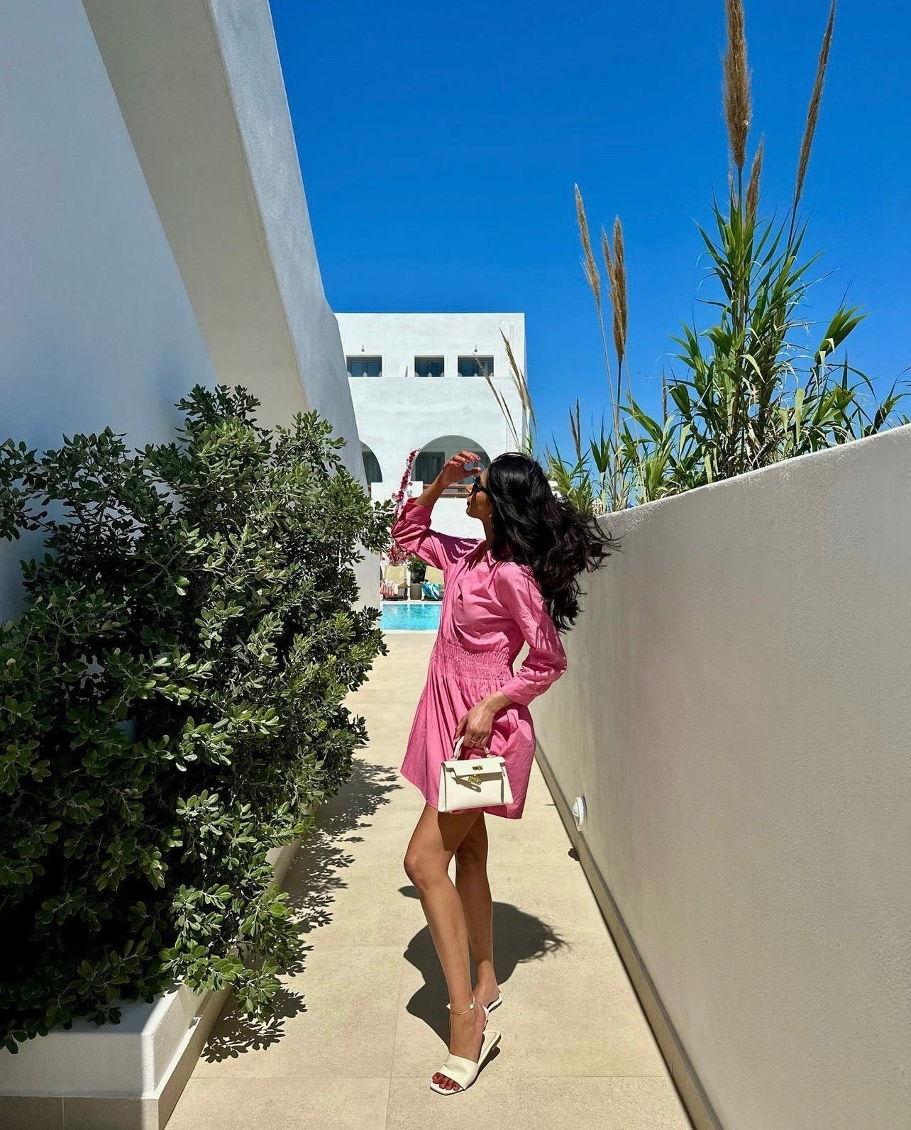 Een vrouw in een roze jurk en witte hakken staat op een zonnig pad, haar ogen afschermend terwijl ze de Liora The Label Aurora Signature Mini Leren Handtas met gouden beslag vasthoudt. Groene planten en witte gebouwen verschijnen onder de helderblauwe hemel.