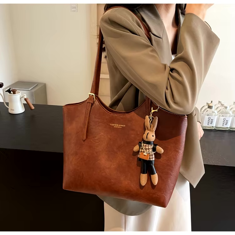 Een vrouw in een beige blazer en witte jurk staat bij een toonbank en houdt de Thalessa Chic Leren Tote Bag met Speelse Charme van Liora The Label vast - een elegante bruine tas met een speelse pluchen konijntje-sleutelhanger voor extra flair.