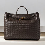 De Alyn Zwarte Geweven Midi Leren Tote Bag van Liora The Label heeft een enkel handvat aan de bovenkant, een gevlochten leren detail aan de voorkant en een gouden knoop accent, weergegeven op een licht oppervlak met een neutrale achtergrond voor tijdloze elegantie.