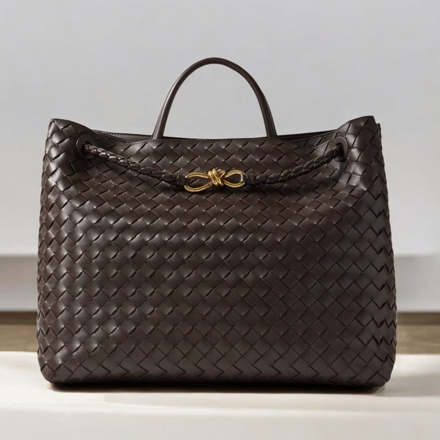 De Alyn Zwarte Geweven Midi Leren Tote Bag van Liora The Label heeft een enkel handvat aan de bovenkant, een gevlochten leren detail aan de voorkant en een gouden knoop accent, weergegeven op een licht oppervlak met een neutrale achtergrond voor tijdloze elegantie.