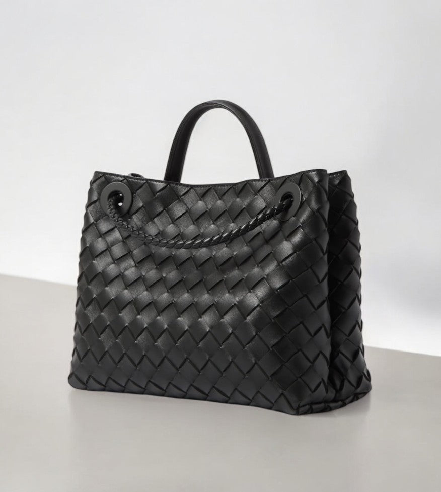 De Alyn Zwarte Geweven Midi Leren Tote Bag van Liora The Label heeft luxe geweven zwart leer, twee korte hengsels en gevlochten details, perfect voor een moeiteloos elegante look. Afgebeeld op een lichte ondergrond tegen een effen achtergrond.