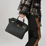 Een persoon in een zwart-bruine outfit en een geruit jasje houdt de Isla, een klassieke zwarte leren schoudertas met gouden sluiting van Liora The Label vast. De achtergrond is effen en lichtgekleurd.