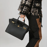 Een persoon in een zwart-bruine outfit en een geruit jasje houdt de Isla, een klassieke zwarte leren schoudertas met gouden sluiting van Liora The Label vast. De achtergrond is effen en lichtgekleurd.