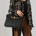 Een persoon in een zwart met beige structuur jasje en bruine broek houdt de Isla vast, een klassieke zwarte leren schoudertas met gouden sluiting van Liora The Label, tegen een effen lichte achtergrond.