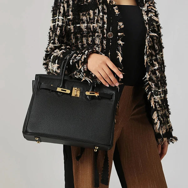 Een persoon in een zwart met beige structuur jasje en bruine broek houdt de Isla vast, een klassieke zwarte leren schoudertas met gouden sluiting van Liora The Label, tegen een effen lichte achtergrond.