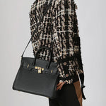 Een persoon draagt een zwart-beige jasje met textuur en draagt de Isla Klassieke Zwarte Leren Schoudertas van Liora The Label, een elegante werkhandtas met gouden sluiting, gestileerd crossbody. De zwarte tas steekt mooi af tegen de neutrale achtergrond.