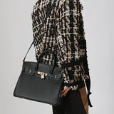 Een persoon draagt een zwart-beige jasje met textuur en draagt de Isla Klassieke Zwarte Leren Schoudertas van Liora The Label, een elegante werkhandtas met gouden sluiting, gestileerd crossbody. De zwarte tas steekt mooi af tegen de neutrale achtergrond.