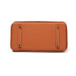 De afbeelding toont de onderkant van de Isla Tan Textured Leather Handtas van Liora The Label, met de rechthoekige vorm, kiezelafwerking en twee parallelle leren stroken met metalen voetjes voor bescherming.