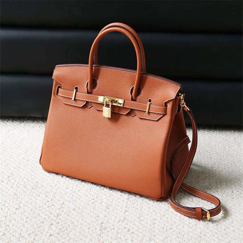 De Isla | Tan Textured Leather Handtas van Liora The Label heeft goudkleurige hardware, een slot, dubbele handgrepen en een afneembare schouderriem, afgebeeld op een licht tapijt met een donkere achtergrond-klassiek & tijdloos elegant.