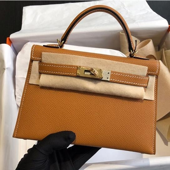 Een hand in een zwarte handschoen houdt de Galatea Elegante Zwarte Leren Handtas van Liora The Label vast, met gouden hardware en gedeeltelijk verpakt in beschermend vilt, in een open doos met wit vloeipapier.