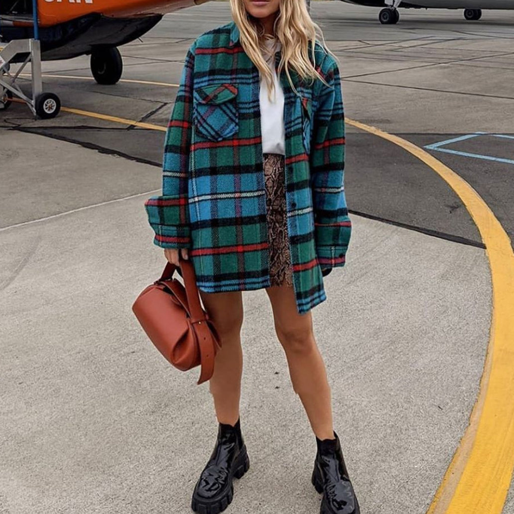 Een vrouw staat op het tarmac van een vliegveld in een geruit overhemd, witte top, rok met patroon, zwarte enkellaarsjes en houdt de Guinevere Bruine Leren Schoudertas van Liora The Label vast. Haar golvende haar bedekt haar gezicht gedeeltelijk.