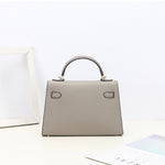 De Aurora Signature Mini Leren Handtas van Liora The Label, een gestructureerde lichtgrijze handtas met gouden hardware, staat op een wit oppervlak bij een fotolijst en tijdschrift tegen een effen witte achtergrond.