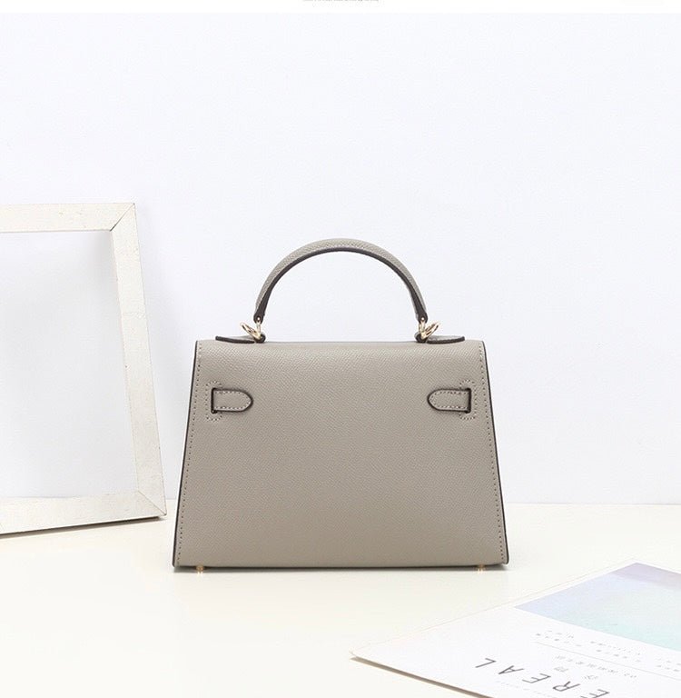 De Aurora Signature Mini Leren Handtas van Liora The Label, een gestructureerde lichtgrijze handtas met gouden hardware, staat op een wit oppervlak bij een fotolijst en tijdschrift tegen een effen witte achtergrond.