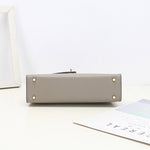 De Aurora Signature Mini Leren Handtas van Liora The Label in het grijs ligt plat op een wit oppervlak, de basis met vier gouden noppen zichtbaar. Vlakbij liggen een wit fotolijstje en een minimalistisch tijdschrift op tafel.