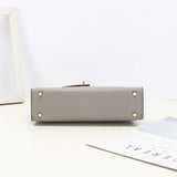 De Aurora Signature Mini Leren Handtas van Liora The Label in het grijs ligt plat op een wit oppervlak, de basis met vier gouden noppen zichtbaar. Vlakbij liggen een wit fotolijstje en een minimalistisch tijdschrift op tafel.