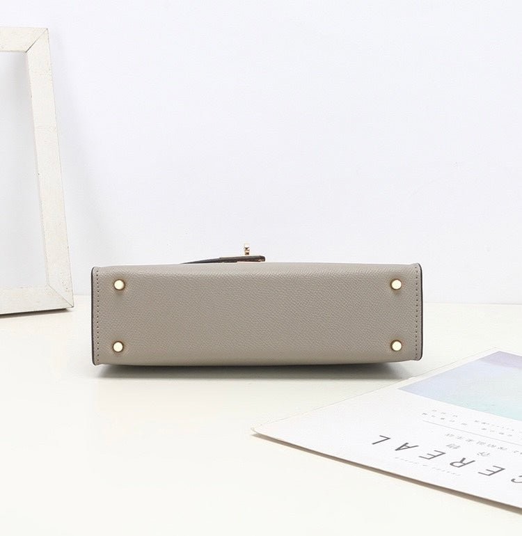 De Aurora Signature Mini Leren Handtas van Liora The Label in het grijs ligt plat op een wit oppervlak, de basis met vier gouden noppen zichtbaar. Vlakbij liggen een wit fotolijstje en een minimalistisch tijdschrift op tafel.